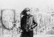 A Black man in a flat cap and jacket stands central against a graffiti'd wall, staring directly at the camera.